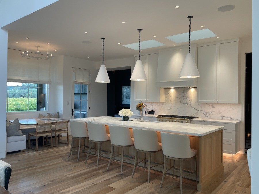 A modern kitchen and living area with windows showcasing a scenic forest view. Features a cozy dining nook, elegant pendant lighting, recessed lighting, and a large kitchen island.
