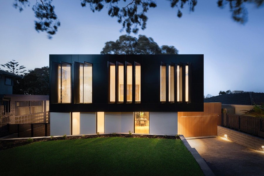Modern two-story house with tall vertical windows glowing at dusk, featuring a sleek black and white exterior and a well-lit entryway.