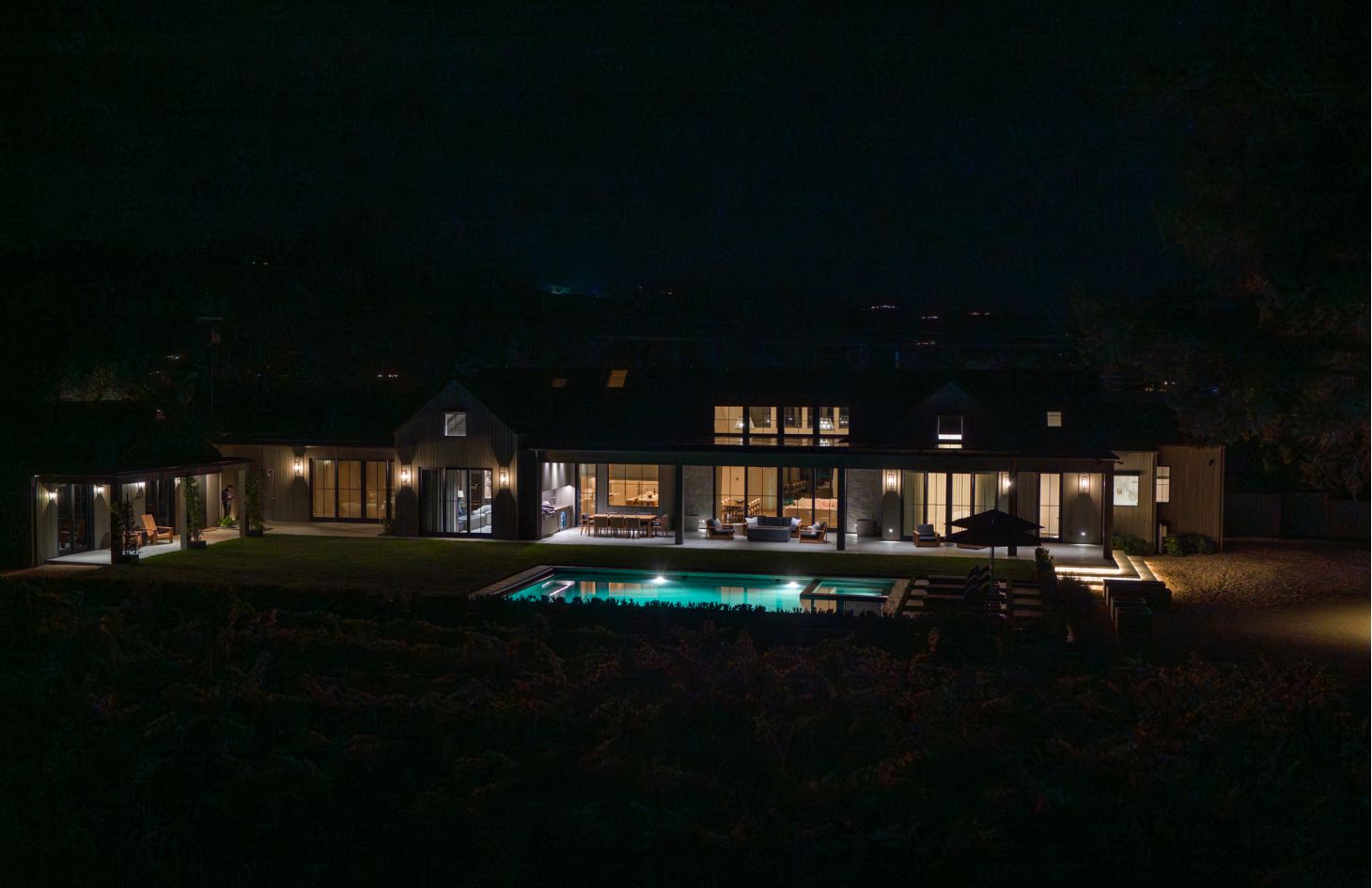 Rear elevation at night; interior rooms glow through tall windows while pool and terrace lights reflect off the water against a dark hillside.
