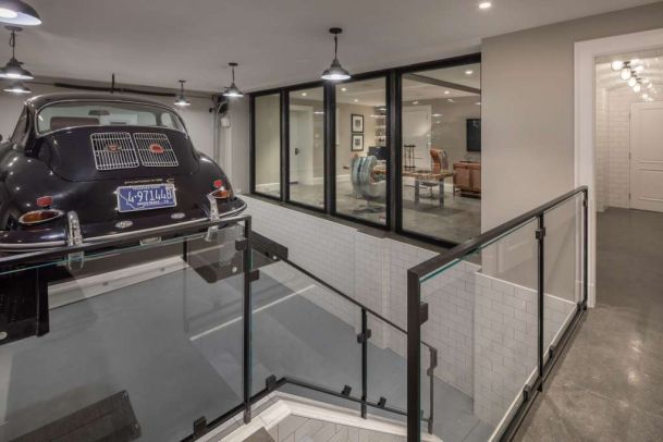 Modern home interior with a classic black car parked on an upper garage level beside a glass-railed stairwell, looking through large interior windows into a stylish lounge area below.