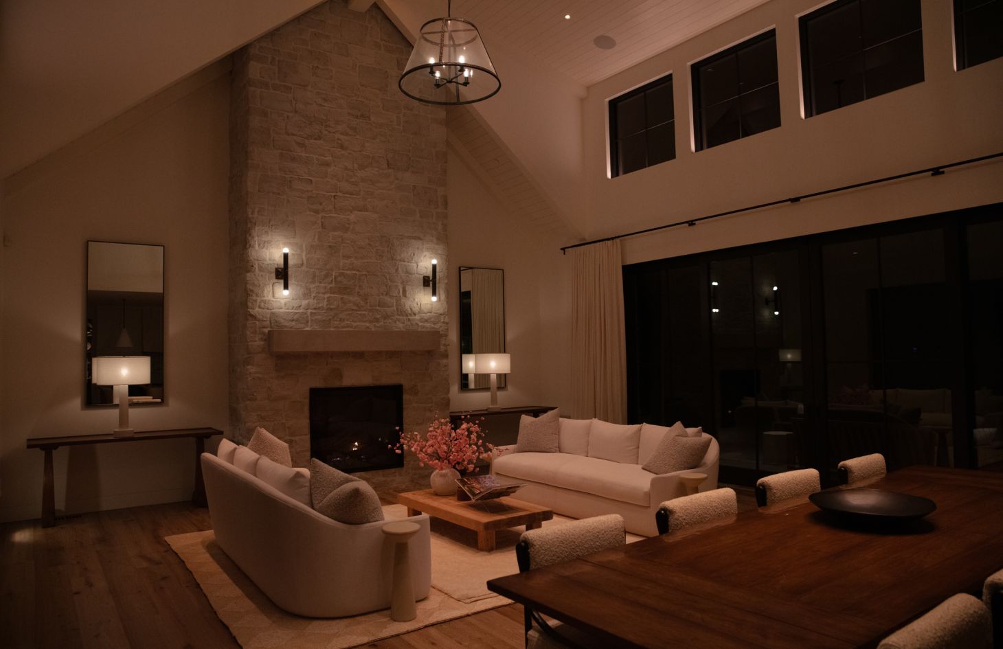 Cozy living-dining space with cathedral ceiling; stone fireplace and matching mirrors anchor the room, while neutral sofas, a wood coffee table, and warm accent lighting create an inviting ambience.
