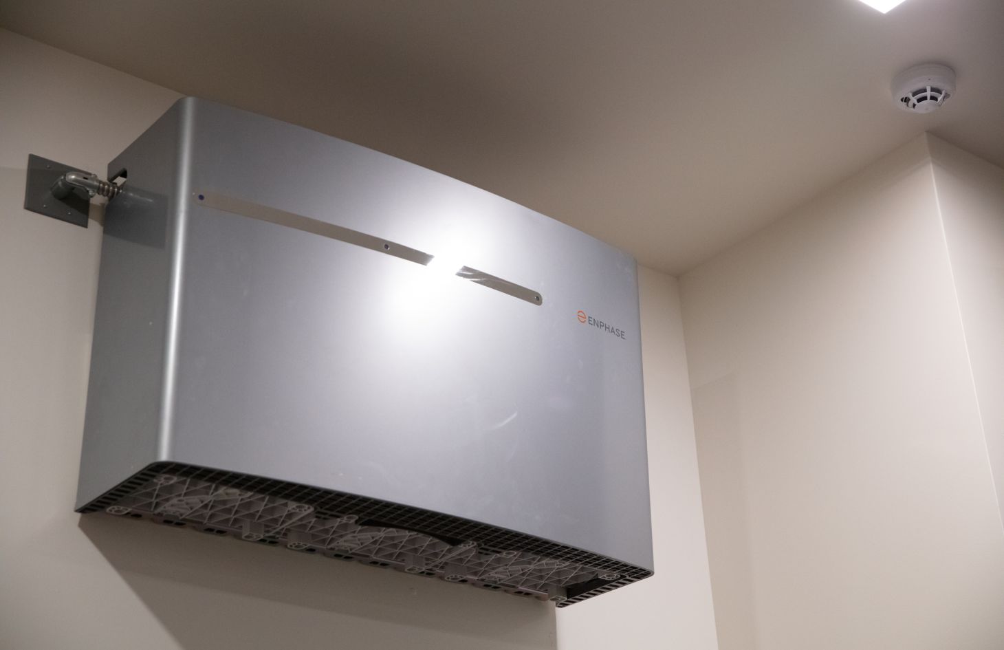 Low-angle close-up of a single gray battery unit and conduit bracket against a smooth white garage wall next to a ceiling smoke detector.