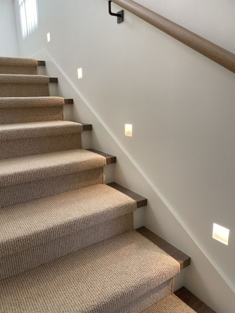Carpeted staircase with wooden step edges and small square wall lights illuminating the steps along a simple handrail.