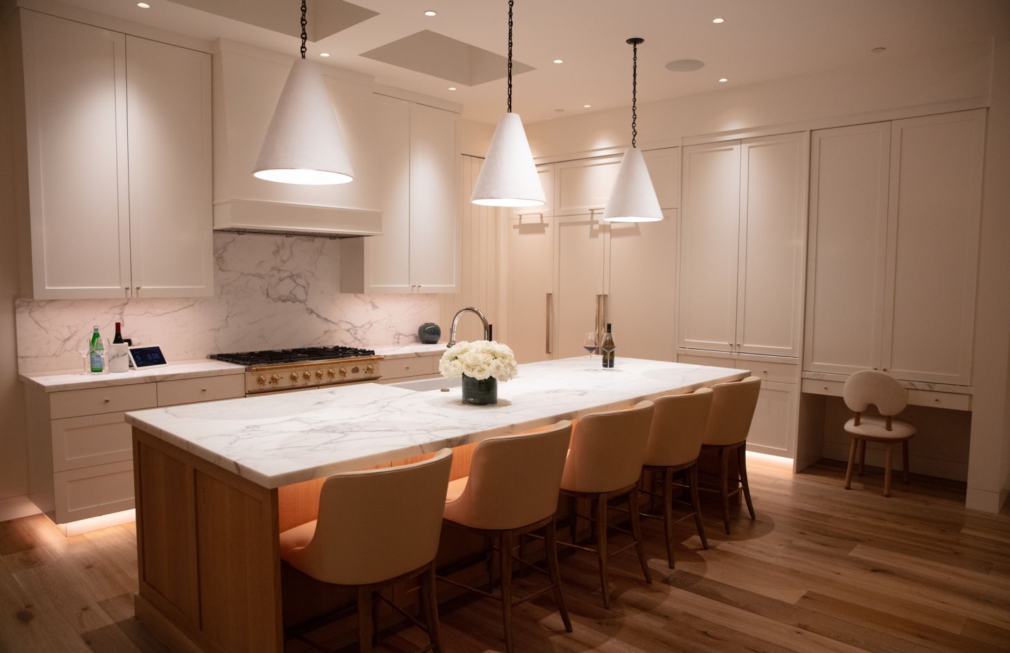 Elegant, well-lit kitchen featuring a large marble waterfall island with five tan barstools, three white cone pendant lights, white shaker cabinetry, and light wood floors.