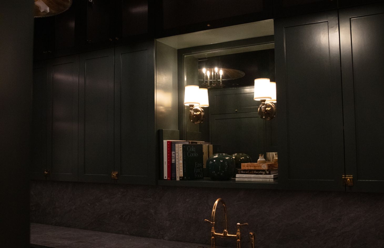 Close view of the pantry’s pass-through niche: twin brass-and-white sconces flank a mirror above cookbooks, pottery, and a matte brass faucet on a dark stone counter.