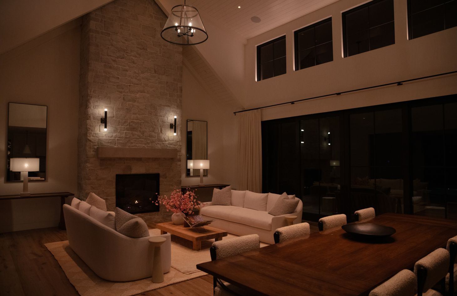 Nighttime view across a dining table into a softly lit living room: stone fireplace, two cream sofas, pink floral centerpiece on a wood coffee table, and layered lighting from sconces, table lamps, and recessed ceiling fixtures.