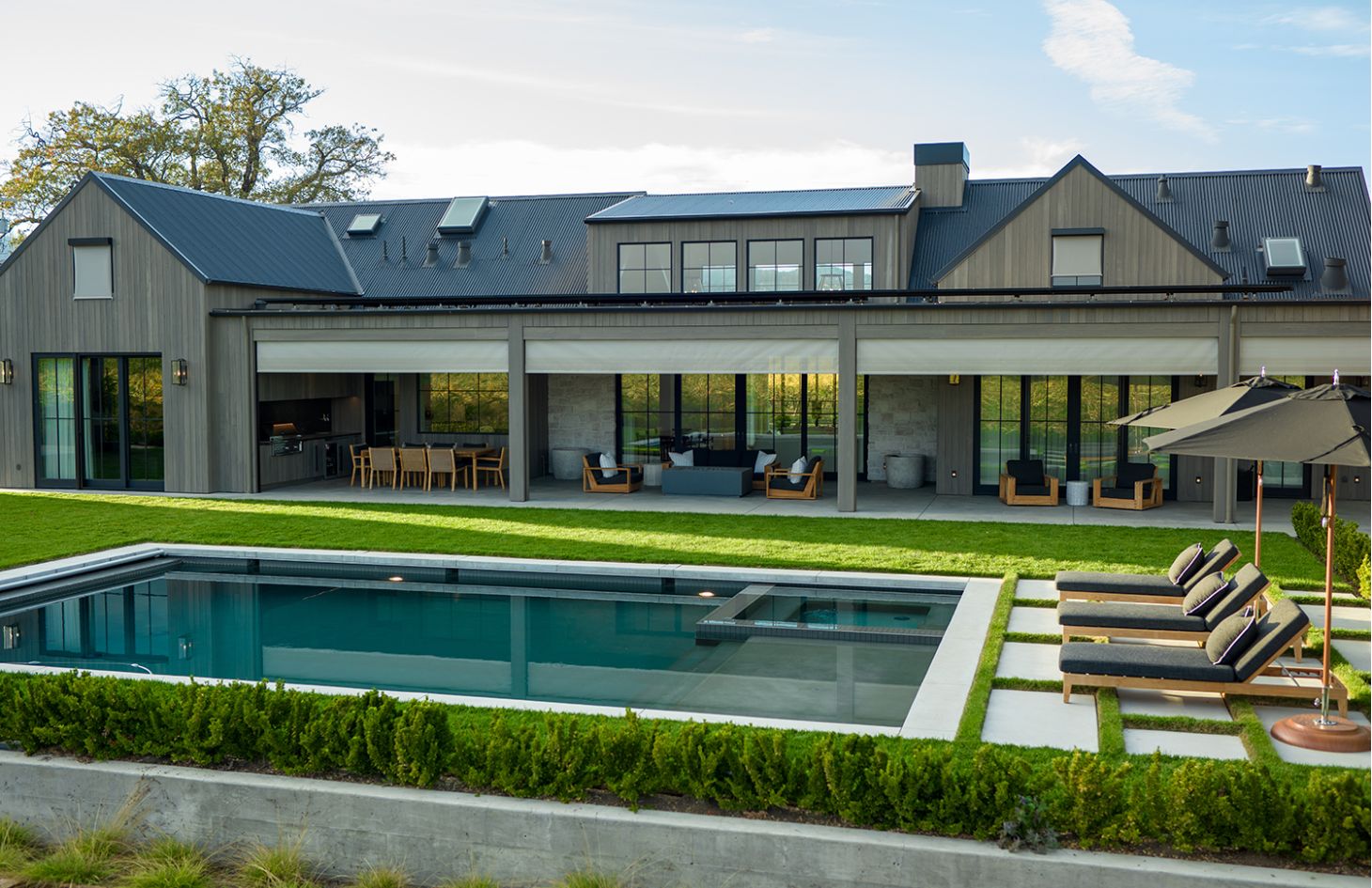 Backyard view of the same house: covered patio with dining and lounge sets opens to a rectangular pool, manicured lawn, and four chaise lounges under black umbrellas.