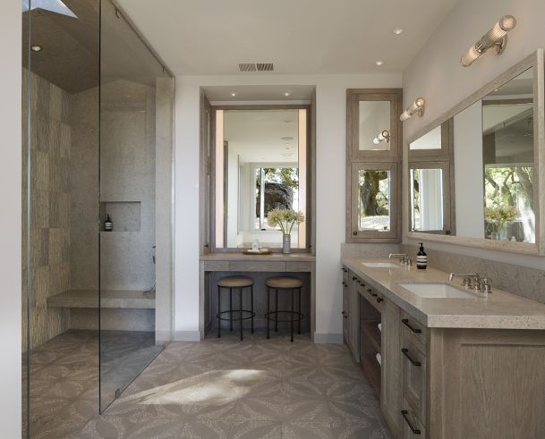 A modern luxury bathroom with a glass walk-in shower, double vanity, large mirrors, and a built-in makeup area with stools.