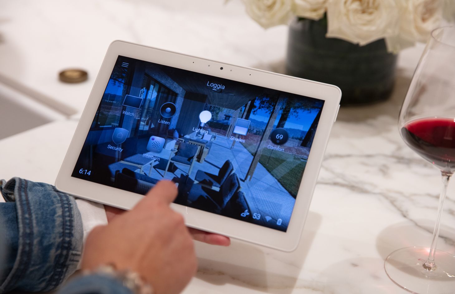 Hand operating a white touchscreen tablet displaying a smart-home control interface on a marble island, with a glass of red wine and bouquet of white roses nearby.