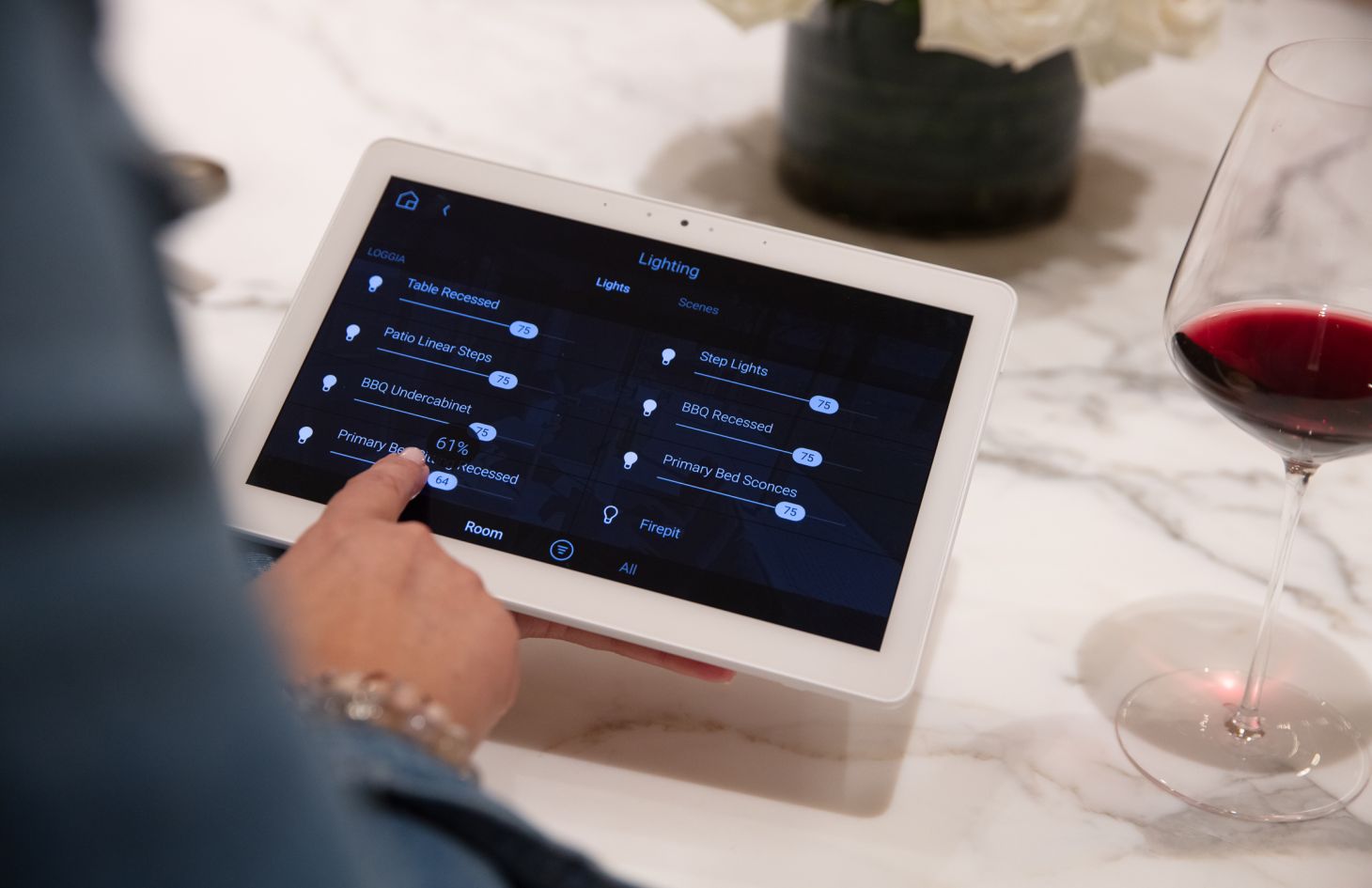 Person adjusting lighting levels on a white touchscreen tablet atop a marble island, with a glass of red wine and white-rose centerpiece nearby.