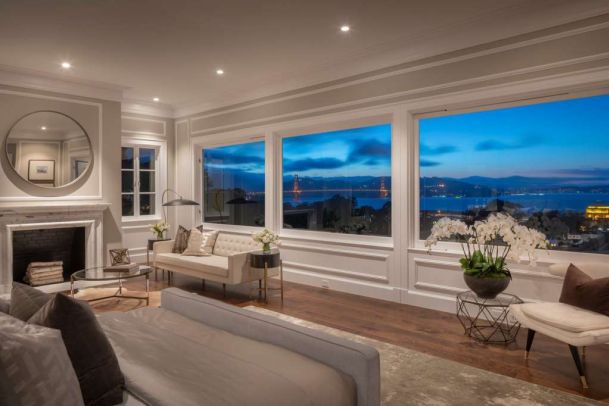 Elegant living room with neutral-toned sofas, fireplace, and hardwood floors, featuring large picture windows that frame an evening view of the bay and a brightly lit suspension bridge.