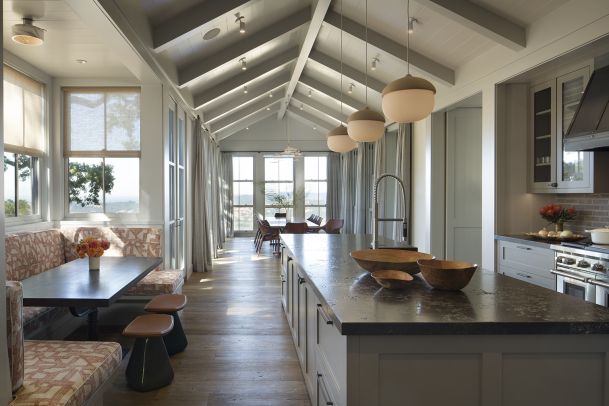 Bright, open kitchen and dining area with a vaulted ceiling, large island and pendant lights, built-in banquette seating on the left, and floor-to-ceiling windows at the far end overlooking an outdoor view.