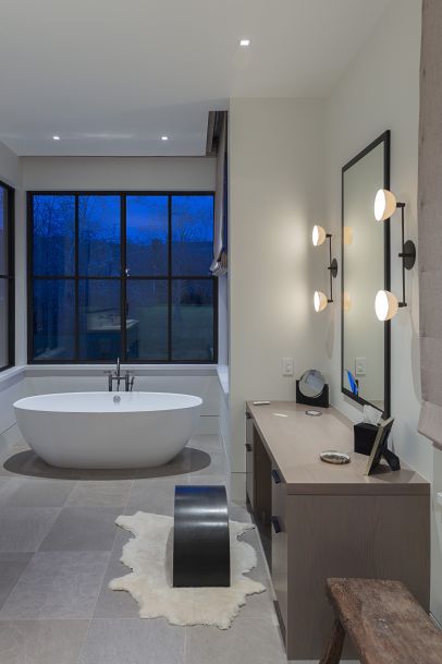 A modern bathroom featuring a freestanding soaking tub by a large window, a vanity with mirror lighting, and contemporary minimalist design elements.