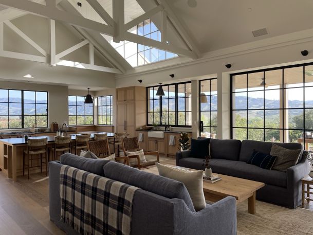 A bright open-concept living room and kitchen with vaulted ceilings, exposed beams, large windows, and scenic mountain views.