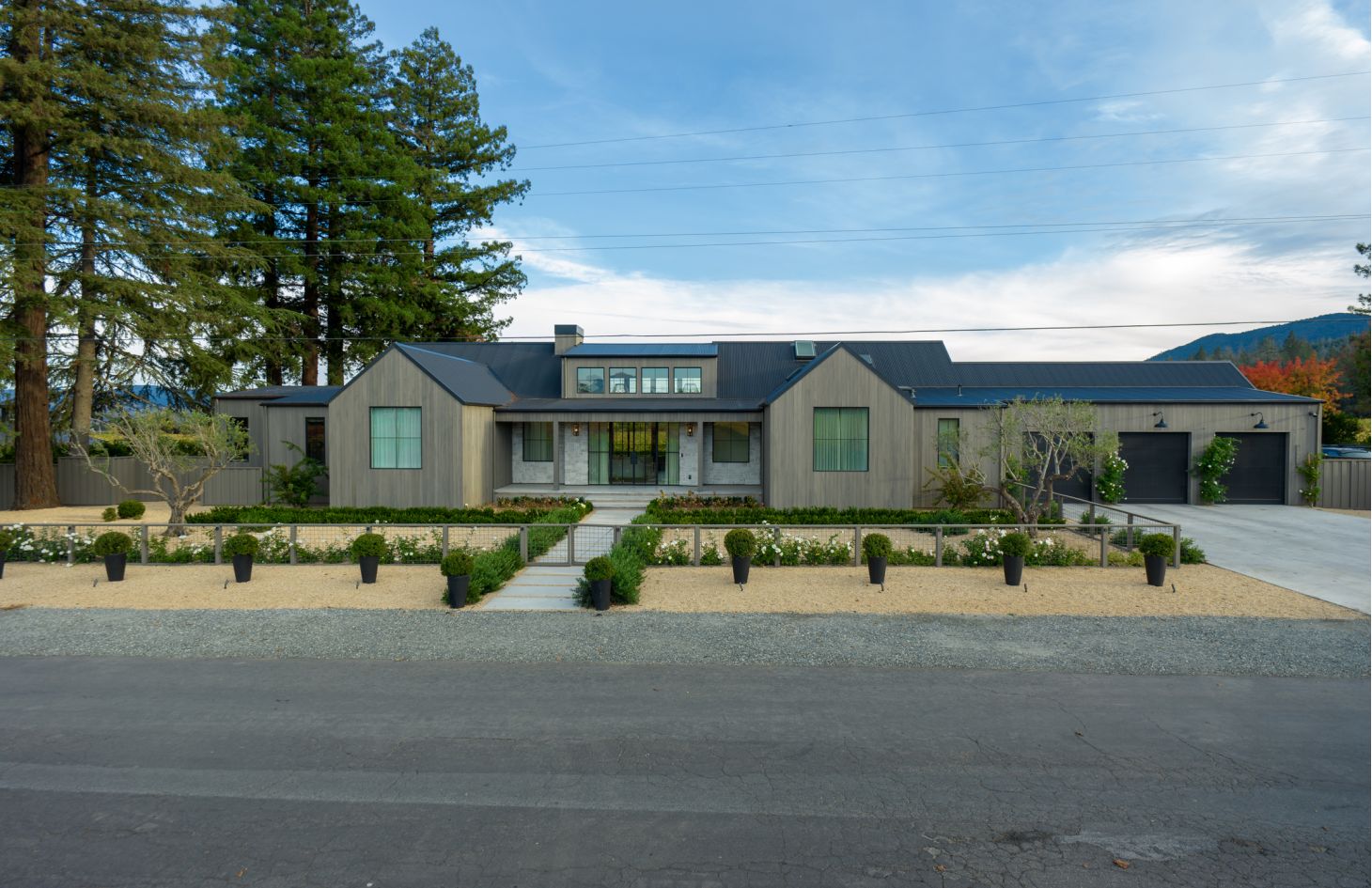 Modern ranch-style home with cedar siding and dark metal roof, framed by tall pines, viewed straight-on behind minimalist gravel beds and clipped hedges.