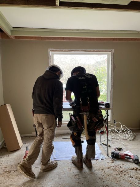 Two construction workers in work clothes and tool belts stand at a table in front of a large window inside a partially renovated room, with tools and extension cords scattered on the unfinished floor.