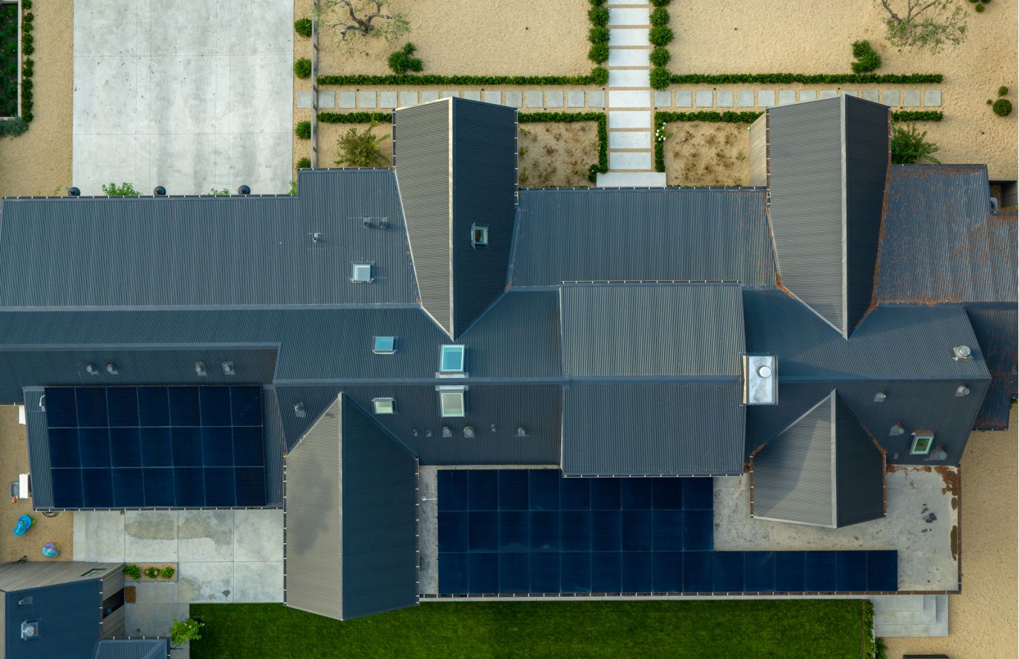 Straight-down drone view of the corrugated metal roof showing skylights, vents, and two large rectangular solar-panel arrays within orderly gravel gardens.