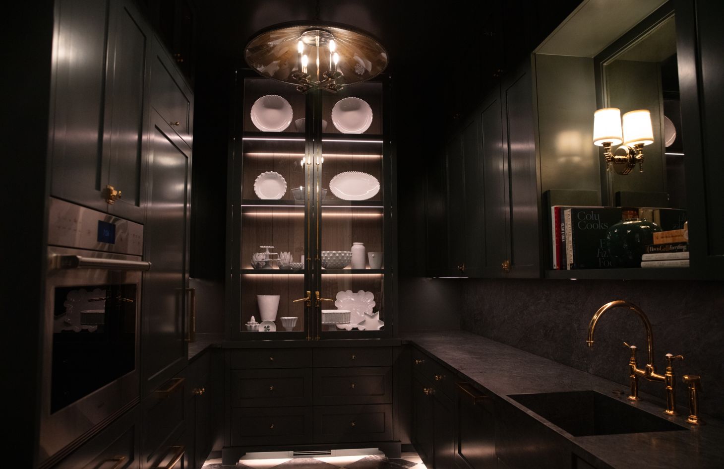 Dramatic butler’s pantry with deep green cabinetry, brass hardware, glass-front display of white china lit from within, stone counters, and a brass gooseneck faucet under warm sconces.
