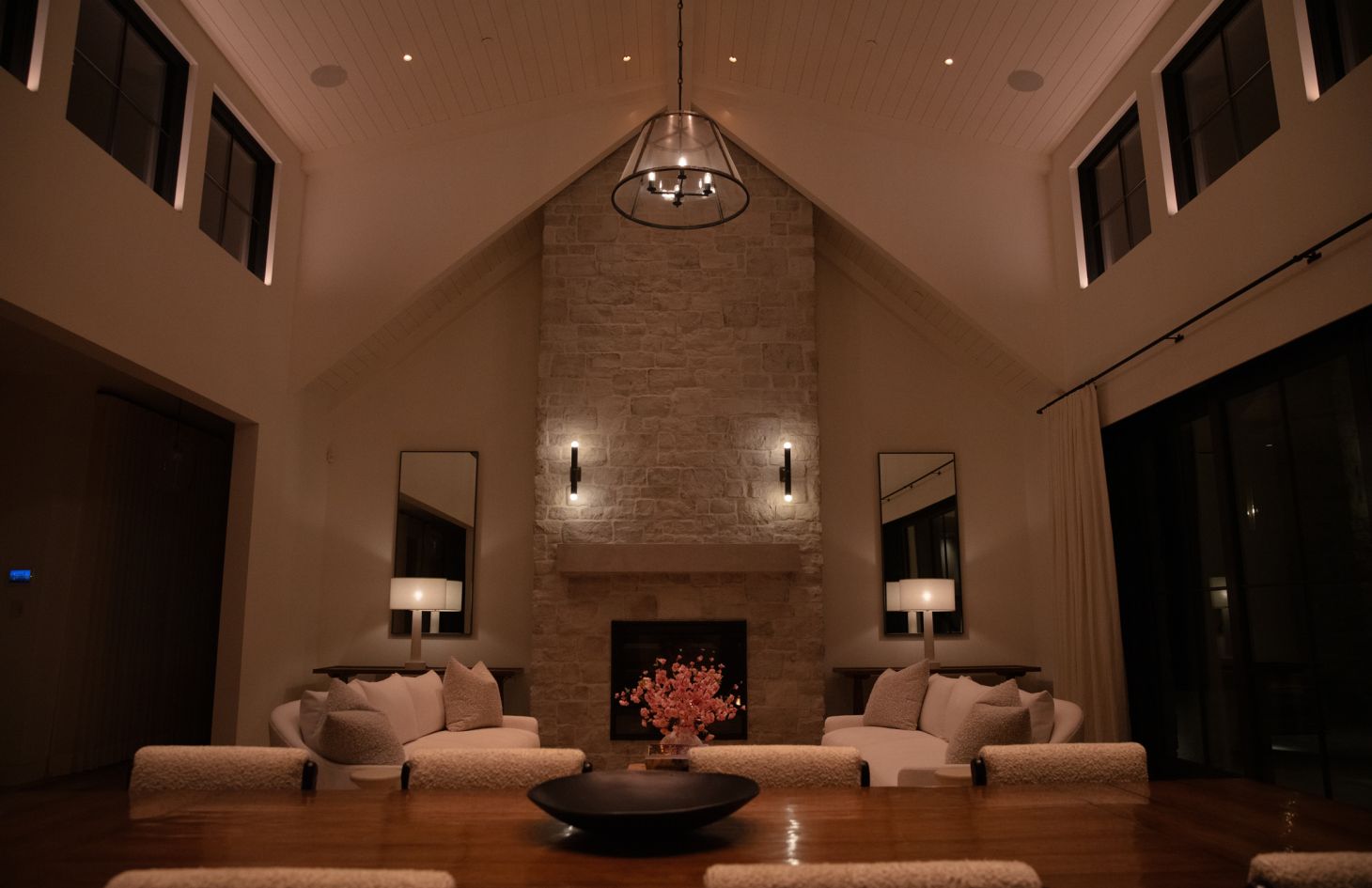 Symmetrical frontal view of a vaulted living room: stone fireplace centered between tall mirrors and table lamps, twin white sofas with textured pillows, and dim uplighting that frames high windows and ceiling beams.