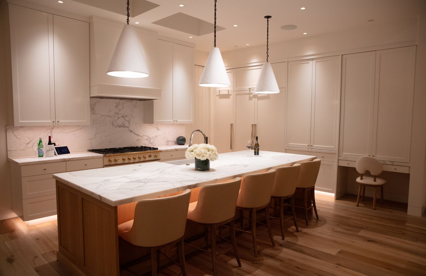 Elegant, well-lit kitchen featuring a large marble waterfall island with five tan barstools, three white cone pendant lights, white shaker cabinetry, and light wood floors.